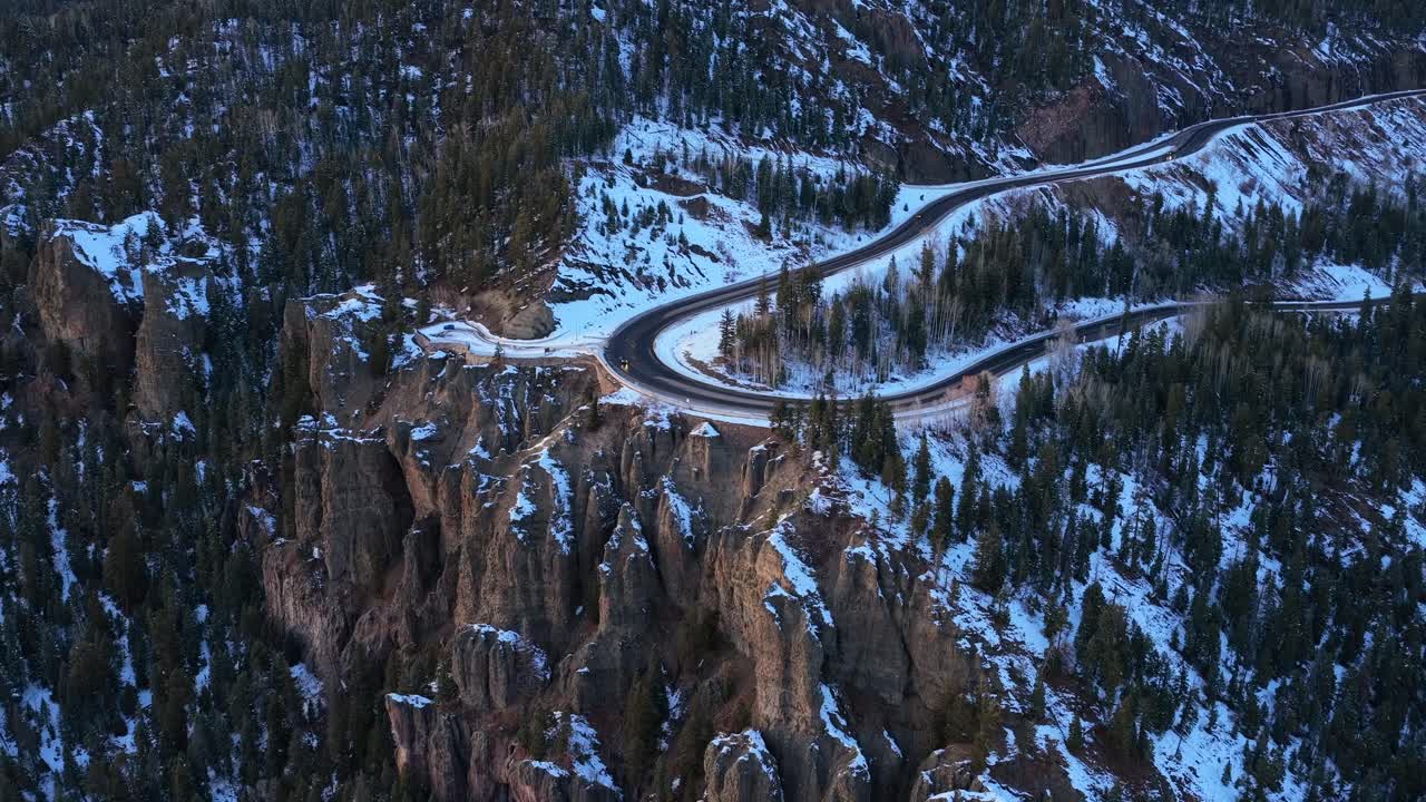 High-angle drone shot of cars driving around a tight hairpin curve on a snowy mountain pass. The winding road cuts through a dense pine forest, highlighting the challenges of winter mountain travel