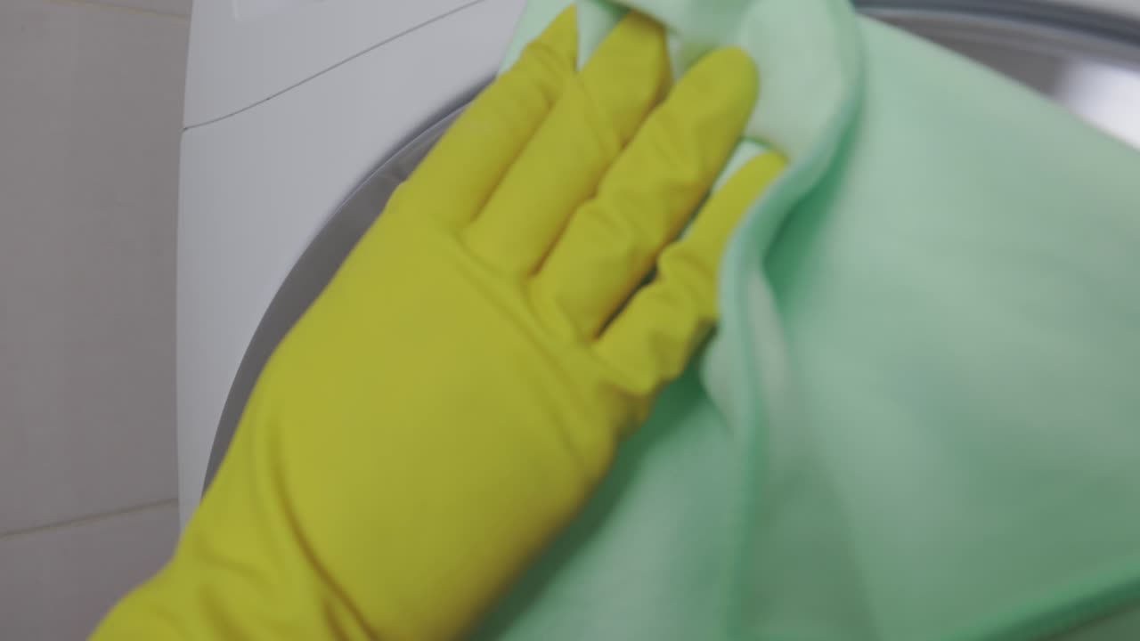 en el baño, un limpiador limpia un tambor de la máquina de lavar de color acero con un trapo verde. la mano de una mujer en un guante de látex amarillo limpia el tambor de una máquina de lajar a un brillo con un trape verde