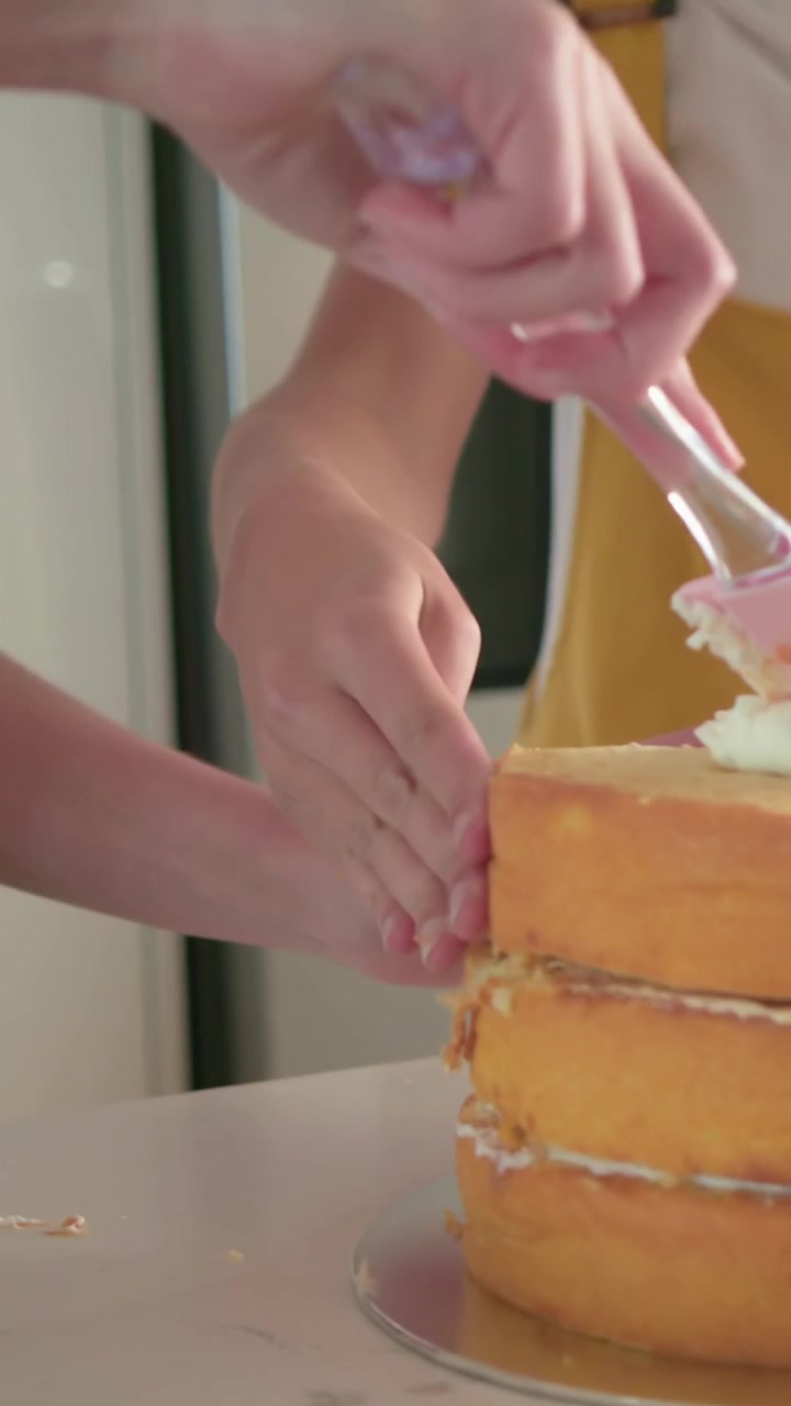 Vertical video: Picking spatula, blonde in pink top icing cake in kitchen, friend steadying