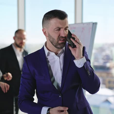 Emotional businessman speaking on the smartphone. Man speaks and gestures actively. Employees standing at the chart in the backdrop