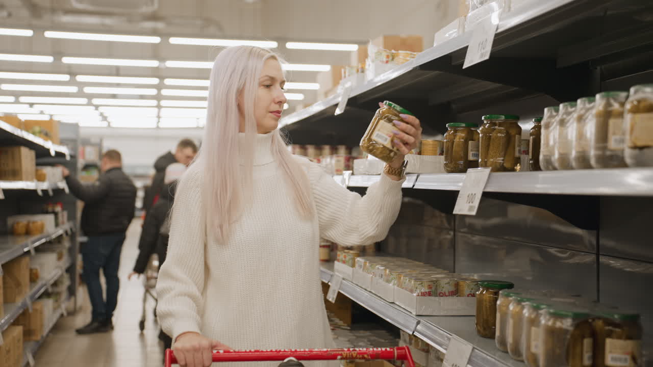 Close up shot showing female shopper pushing cart reaching for glass jar of pickled cucumbers on supermarket shelf lined with jars and cans while other customers browse in bright retail aisle