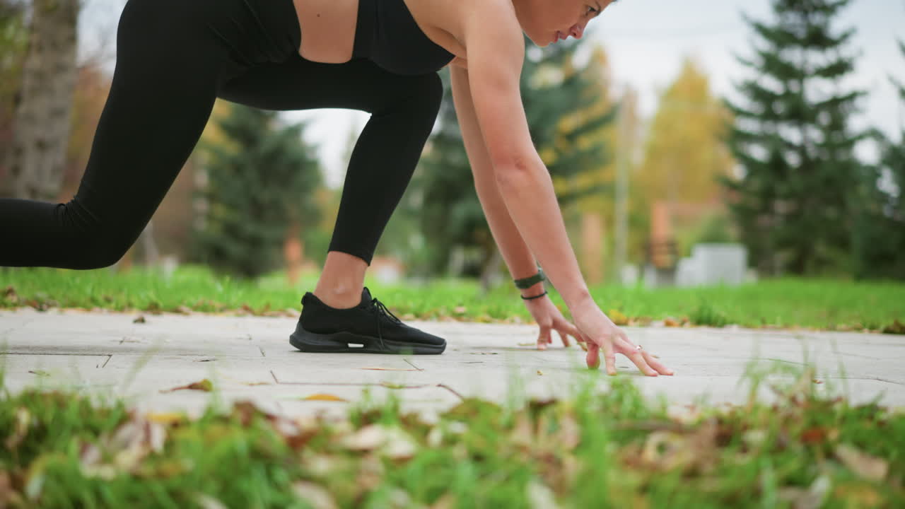 Rotating view of athletic woman in black leggings getting ready to run outdoors, with hands on the ground, preparing for takeoff in park, focusing on her form against background of greenery