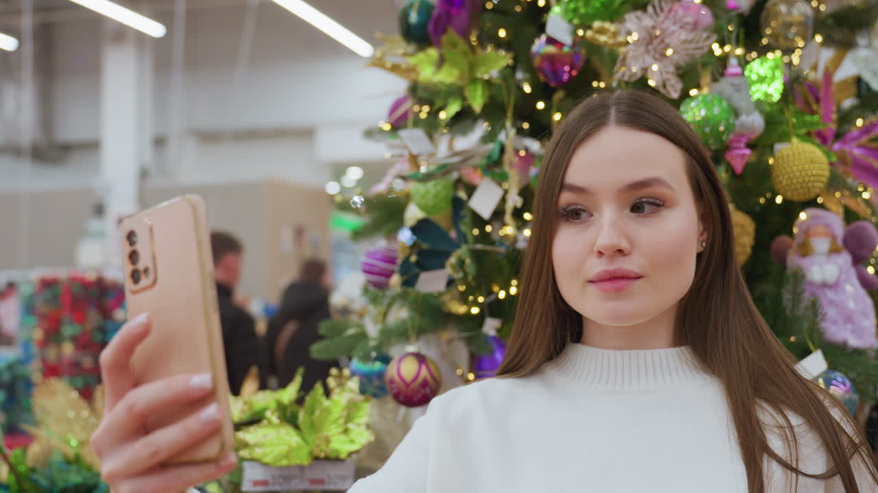 Elegant lady in white sweater takes a selfie in front of vibrant Christmas decor in a well-lit store, she adjusts her hair while a shopper walks by in the background