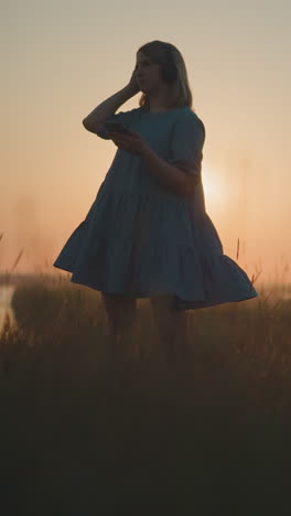 woman in a blue gown, swaying gently, enjoys a sunset while using a smartphone and wearing a headset in a glowing, sunlit field