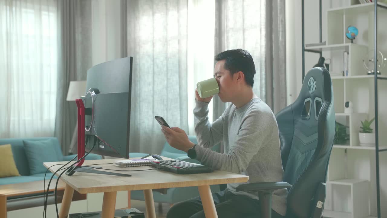Asian Man In Long Sleeved T-Shirt And Black Pants Using Mobile Phone And Drinking Coffee While Using Desktop Computer For Working At Home.