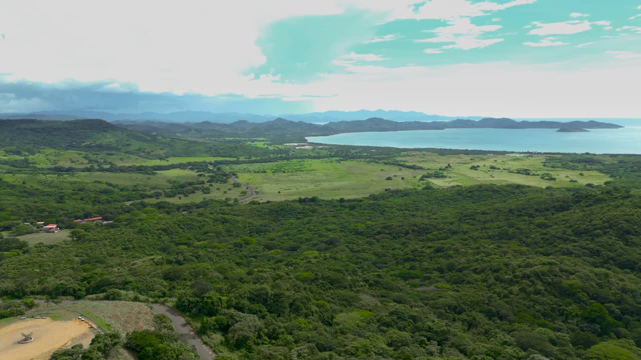 vista aérea de un paisaje que tiene montañas lejanas, con pastos verdes, praderas, árboles y playas durante un día soleado mientras los pájaros vuelan a través de la toma