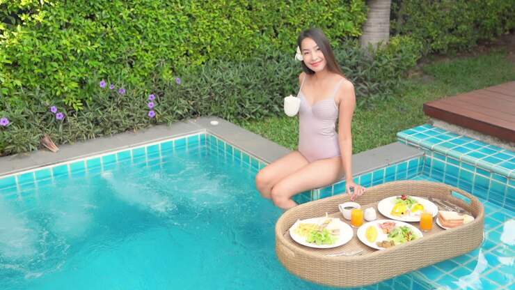 Hight angle, Breakfast is served via floating platter to lovely Asain woman sitting on the ledge of the pool, drinking a cup of coffee