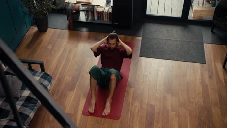 a brunette guy in a red t-shirt and green shorts does an exercise to strengthen the abdominal muscles on a special mat in a country industrial house