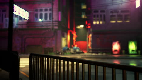 a nighttime view of a city street with neon lights, fog and a metal fence in the foreground.