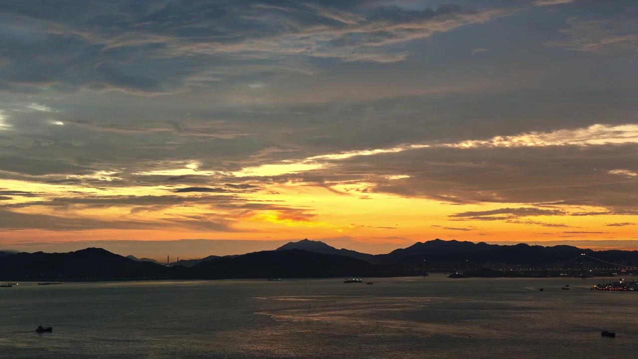 Stunning time-lapse of glowing orange and yellow clouds moving over a calm harbor bay with distant mountains and city lights.