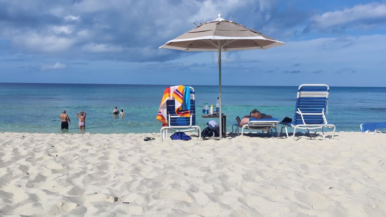 Persone in mare che si godono una calda giornata estiva sull'isola Blue Lagoon alle Bahamas con lettino e ombrellone, ripresa a mano libera da dietro, 4K 30 FPS
