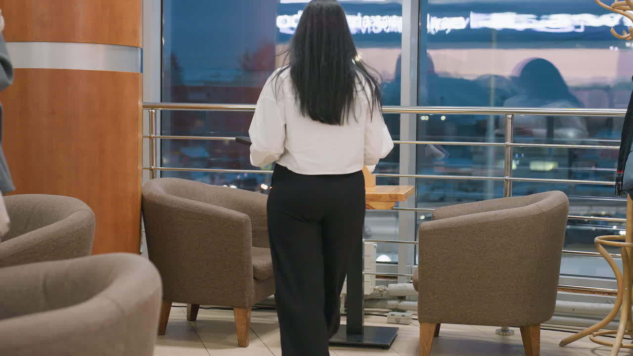 Young woman in white shirt and black pants walks toward indoor cafeteria table holding coffee cup, preparing to take seat, while another person is partially visible in foreground