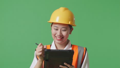 close up of asian female engineer with safety helmet taking note on the tablet and looking around while standing in the green screen background studio