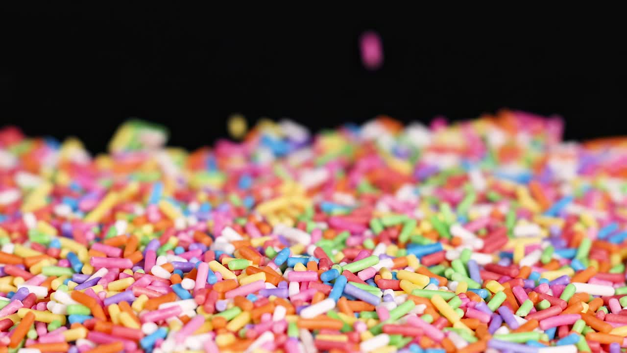 A colorful array of sprinkles accumulates on a dark background, creating a lively and textured scene.