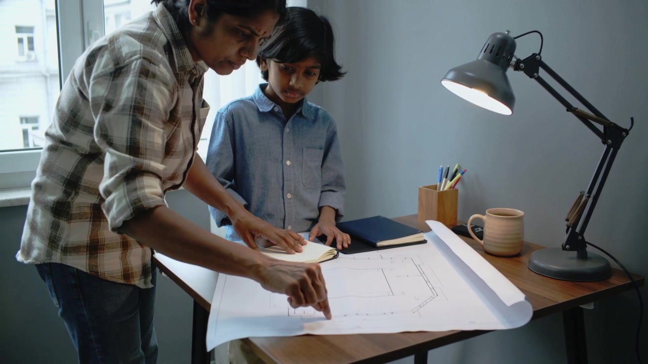 Leaning parent in plaid shirt pointing, tracing blueprint at home desk while son watches with notes