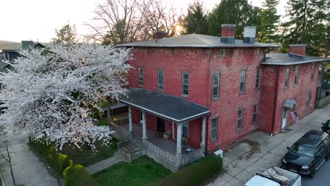 Beautiful-historic-red-brick-house-with-a-large-blooming-tree-in-the-front-yard-and-an-American-flag-gently-swaying-during-sunset