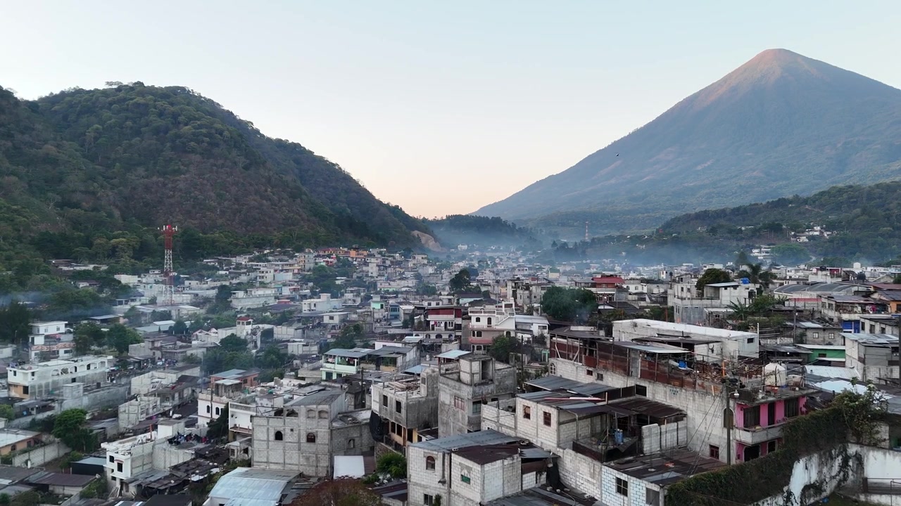 Droneoptagelse over San Lucas, Guatemala, viser tropiske kvarterer, palmetræer, en glødende solnedgangshimmel og skovklædte vulkanbjerge, der omgiver byen i et levende centralamerikansk landskab.