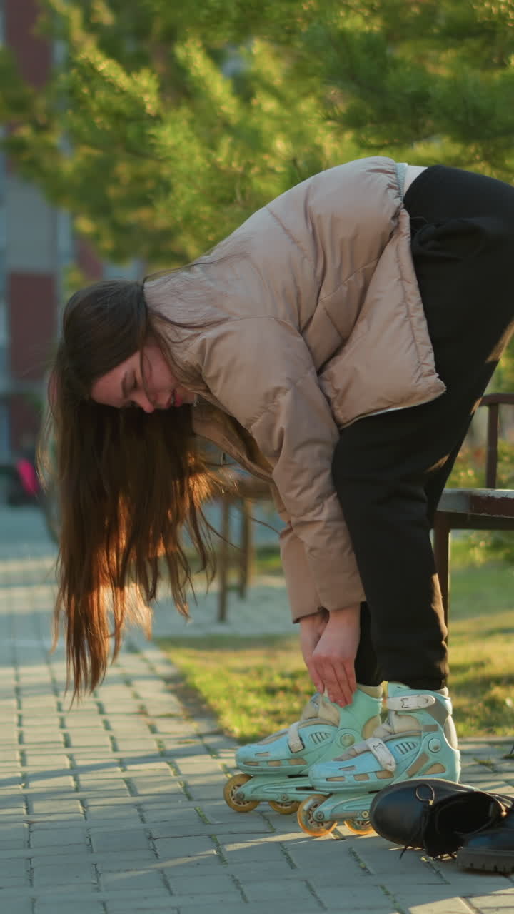 una mujer joven con una chaqueta de melocotón y pantalones negros se inclina para atar sus patines, preparándose para patinar. una bolsa está en el banco a su lado. niños jugando en el fondo
