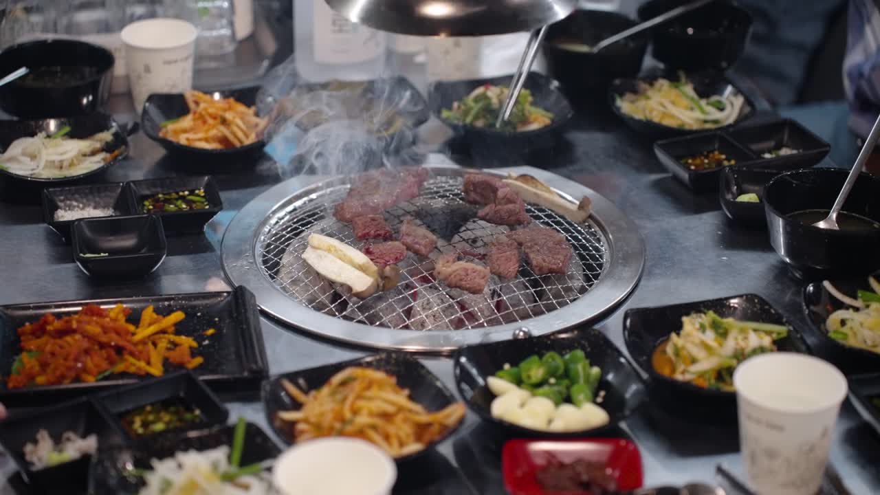 Wide-angle overhead view of hands flipping sizzling Hanu beef and mushrooms on a central table grill, completely surrounded by numerous bowls of colorful, assorted Korean side dishes