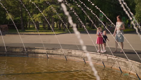 woman with little children walks near fountain in urban park