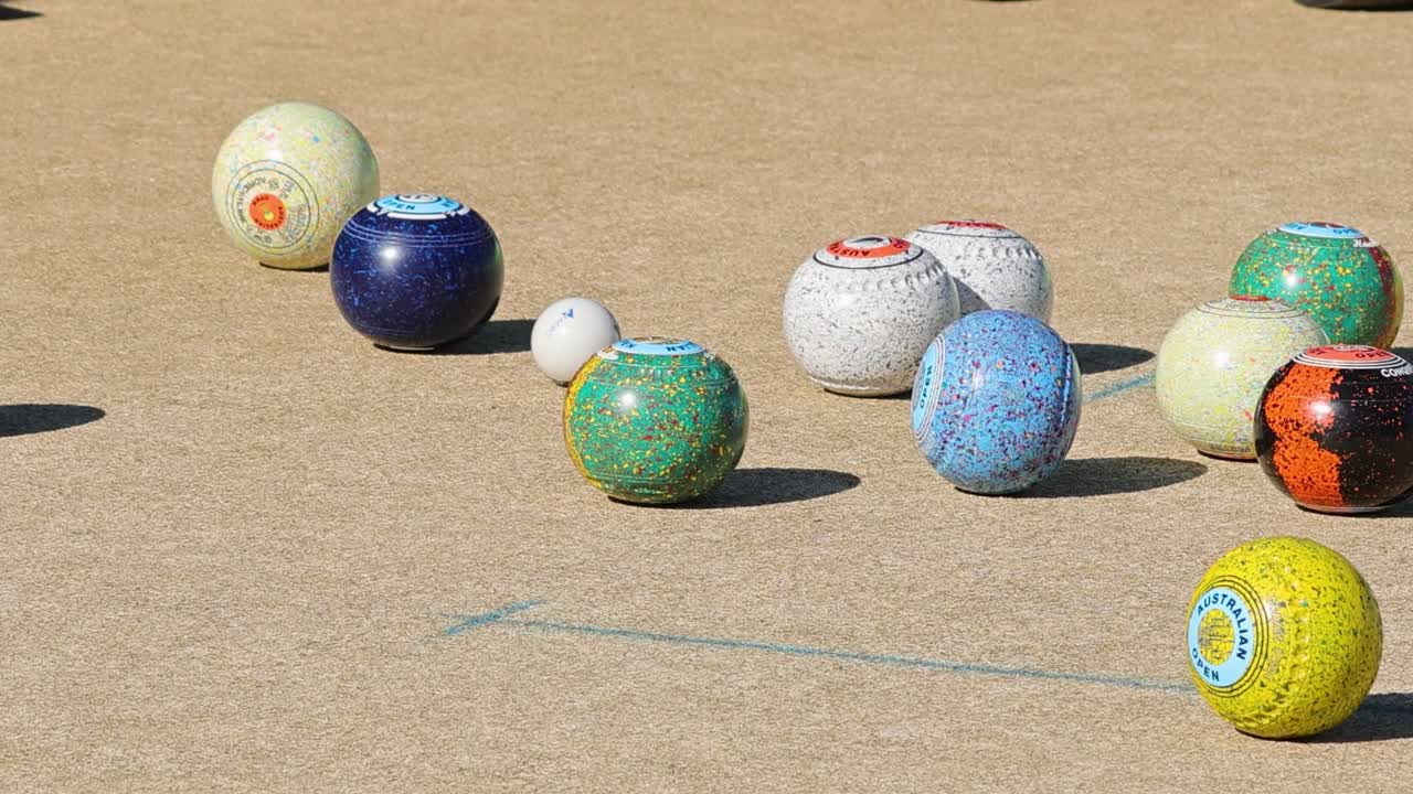 Một bộ sưu tập các quả bóng bowling sân cỏ nhiều màu sắc nằm rải rác trên bề mặt sân bowling cỏ ngoài trời ngập nắng.