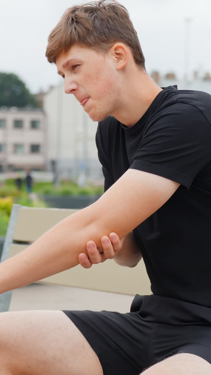 Young caucasian man experiencing acute elbow pain, holding his arm in discomfort, showing an injured area, ache, or health problem outdoors, static vertical slow motion shot