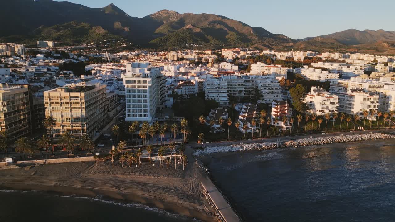 Vista en drone en panorámica izquierda del paseo marítimo bordeado de palmeras de Marbella y del horizonte costero durante un atardecer dorado, con la impresionante costa mediterránea bajo las montañas de Sierra Blanca.