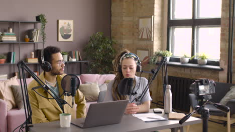 Cucasian-woman-and-african-man-with-headphones-are-sitting-at-table-with-microphone-and-laptop-while-recording-a-podcast