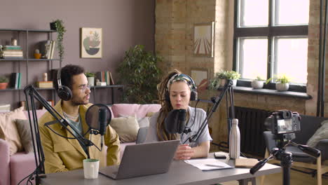 mujer caucásica y hombre africano con auriculares están sentados en la mesa con micrófono y computadora portátil mientras graban un podcast