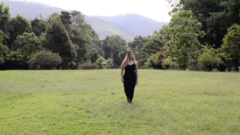 una chica rubia camina sobre una hierba verde y un vestido negro sin zapatos.