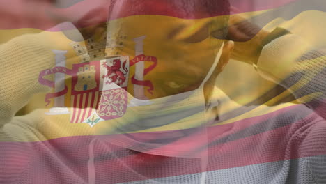 animation of flag of spain waving over man wearing face mask during covid 19 pandemic