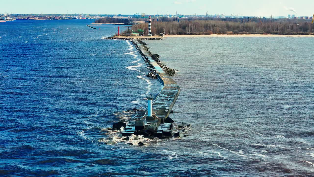 Baltic Sea breakwater in early spring: showcasing a distinct water color divide, with lighthouses guiding the choppy blue and murky gray waters towards a distant forested shore under bright breezy sky