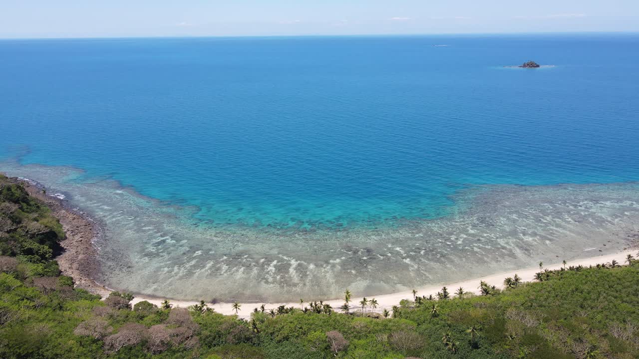 Aerial view of a Fiji island characterized by white sandy beaches, surrounded by clear, shallow waters that transition into deeper blue hues as they extend further from the shore