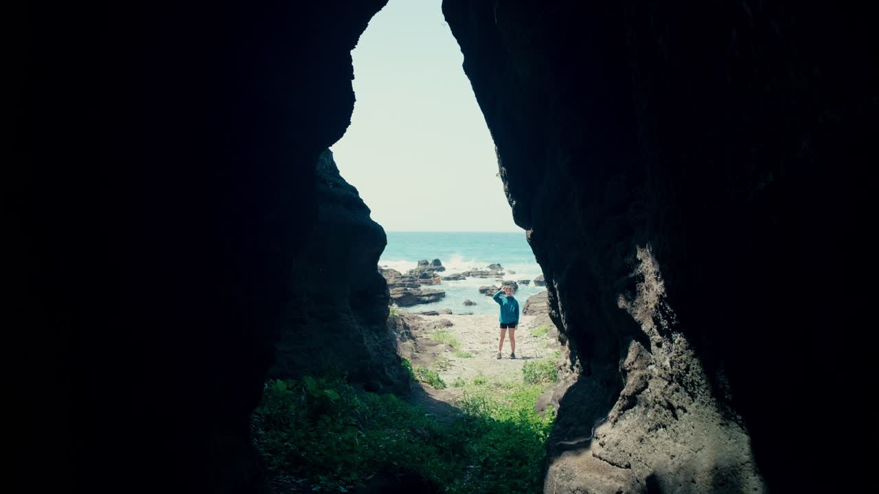 Scenic cave view framing a girl collecting shells on the rocky beach by the sea.