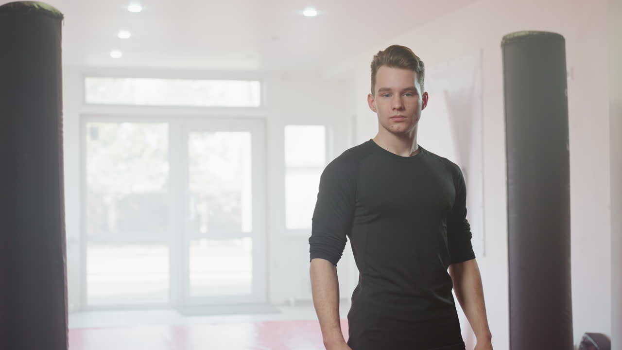 Man in black sportswear stands confidently inside gym between punching bags with serious expression, representing strength, focus, masculinity, determination, discipline, and preparation for intense training