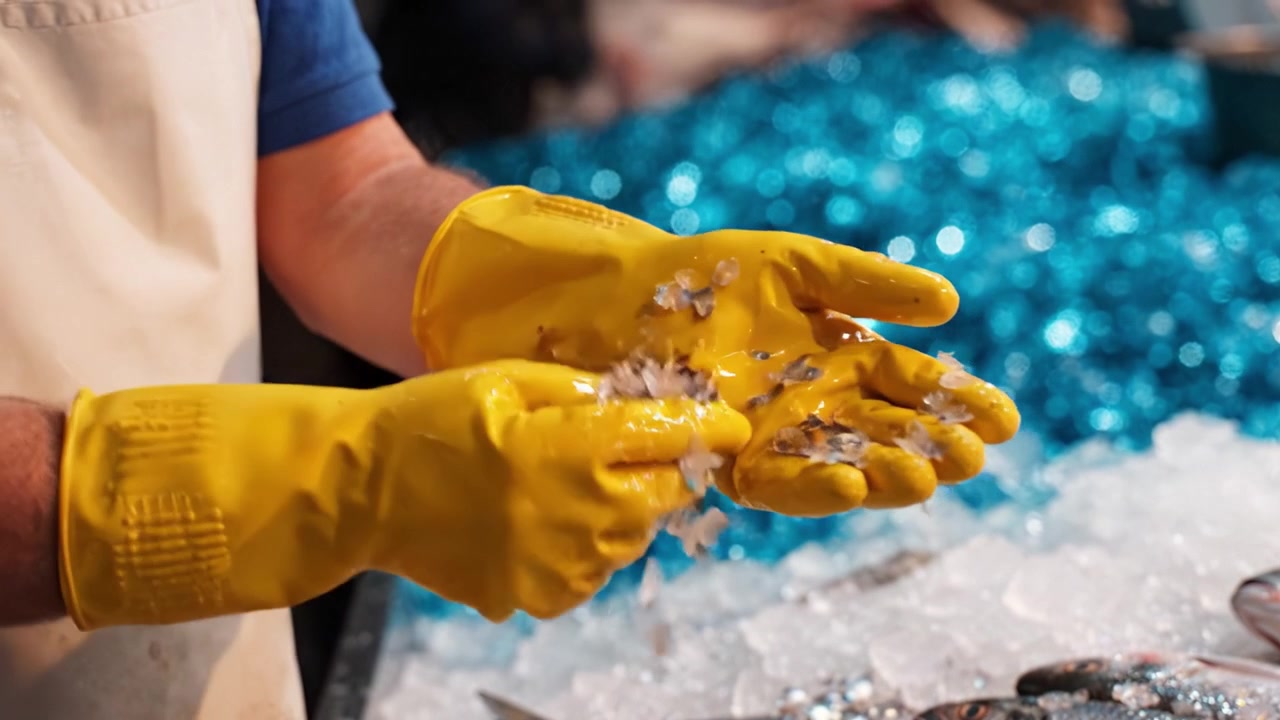 A close-up of a person's hands wearing yellow gloves, expertly cleaning seafood on a bed of ice with sparkling blue decorative stones in the background.
