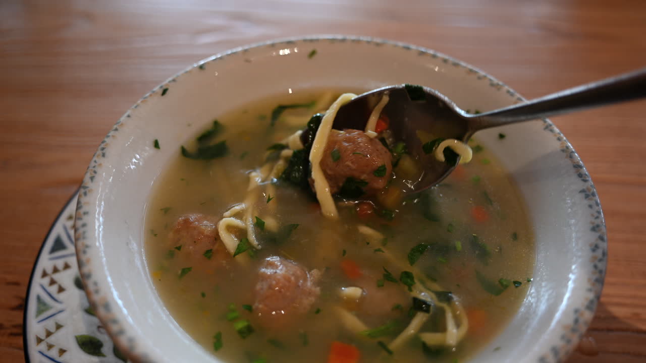 Primo piano di un cucchiaio che solleva noodles e verdure da una zuppa calda e chiara con erbe aromatiche.