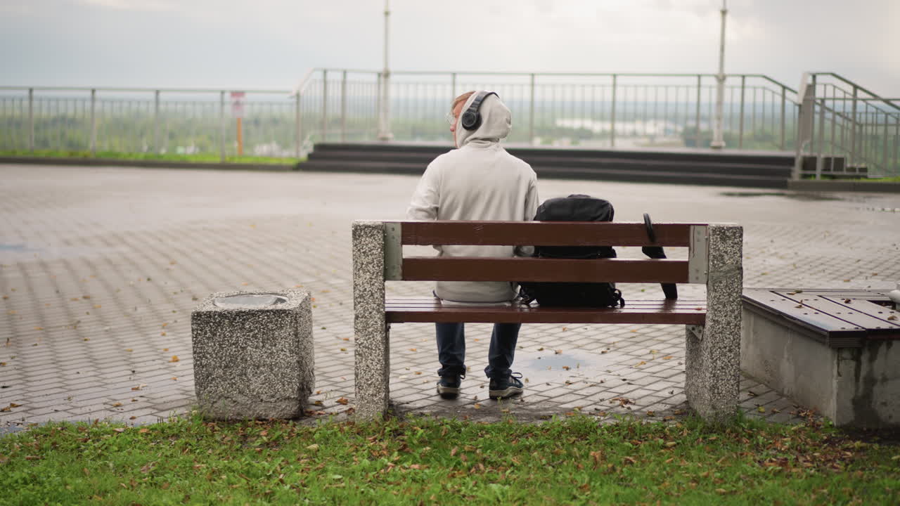 한 남자가 벤치에 앉아 헤드폰으로 음악을 듣고 있어요. 물가, 이슬비가 내리고 젖은 포장도로가 있는 흐린 날씨의 산책로, 좌석 옆에는 작은 여행 가방, 멀리 난간과 수평선이 보이고, 남자가 기다리는 동안 사색에 잠긴 분위기예요.