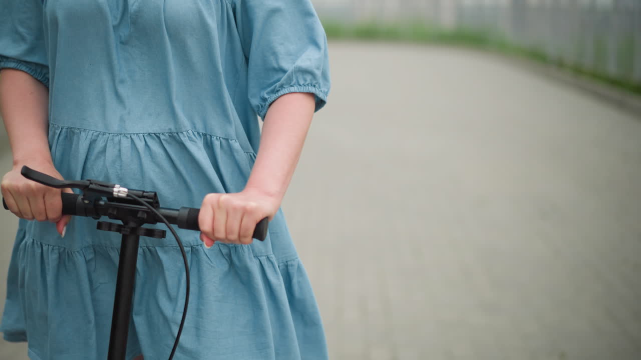 경로에서 스쿠터를 타고 있는 여자의 중간 몸의, 그녀의 파란색 드레스가 바람에 날아다니며 두 손으로 드바를 잡고, 흐릿한 배경으로