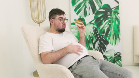 boy touching his belly fat and eating a sandwich sitting on sofa 1