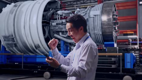 side view of asian male worker with his smartphone with airplane engine maintenance conducted, analytics checking the turbine, he raises his fist up with screaming goal after check on the smartphone