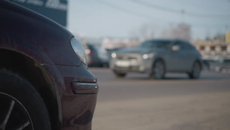 close up maroon car headlight and bumper showing flecks of mud parked on busy urban street with moving cars driving by under bright winter daylight, asphalt pavement and blurred city background