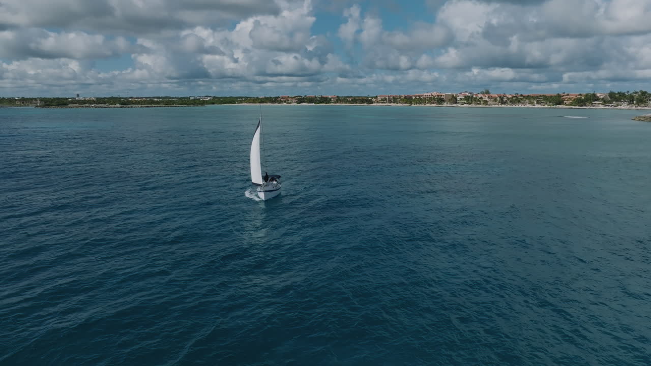 emocionante toma de un avión no tripulado de un yate de vela dejando la costa y aventurándose en el vasto océano, revelando una vista espectacular de la playa circundante, la selva verde y los pintorescos apartamentos de vacaciones