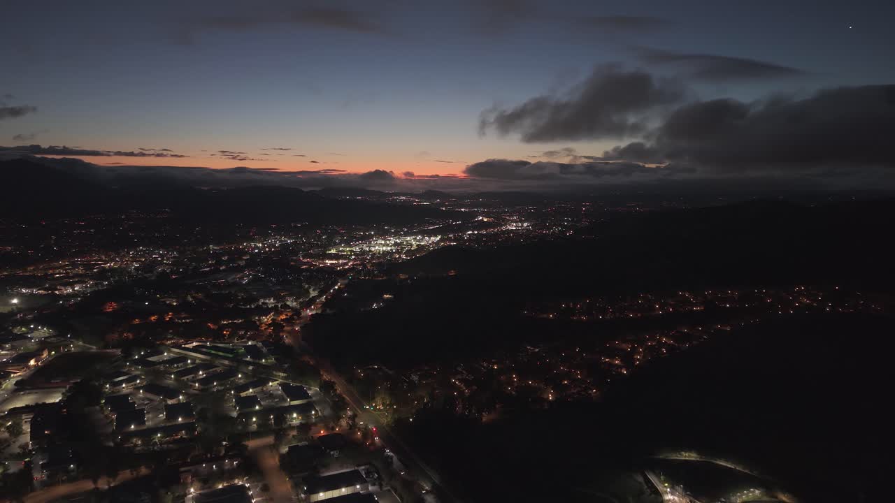Letecký přelet dronem nad Agoura Hills v Kalifornii za soumraku se zářícími městskými světly, dramatickými mraky a obzorem se západem slunce nad předměstskými čtvrtěmi a zvlněnými kopci v malebné večerní atmosféře.