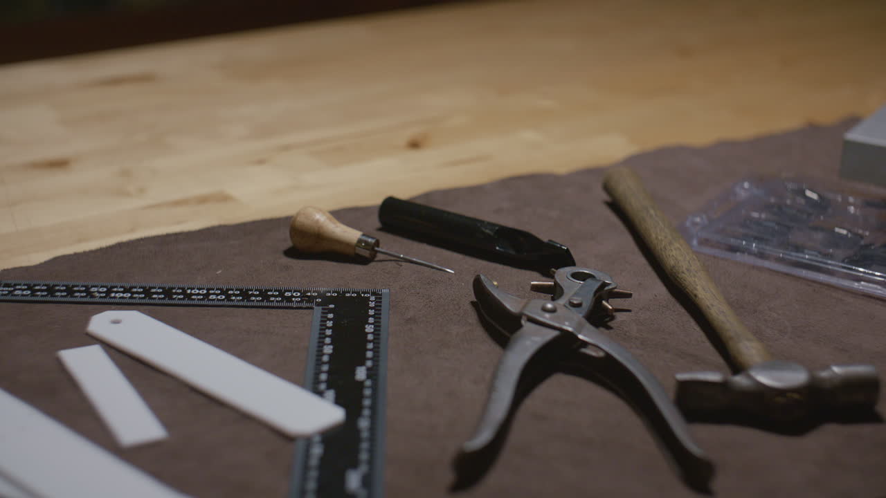 A collection of leather crafting tools, including a punch, awl, ruler, and hammer, neatly arranged on a workbench covered with brown leather.