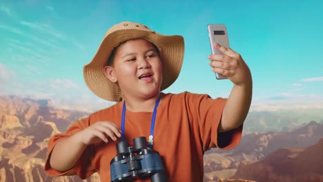 asian boy with a hat and binoculars waving hand having video call on smartphone while traveling at the top of mountain. boy researcher examines something, travel adventure, close up