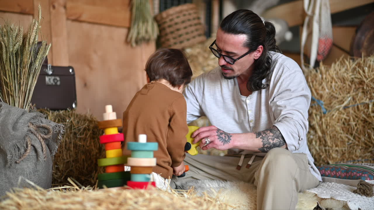 Father playing with his son with colourful, ecological wooden toys in a barn, near square hay bales