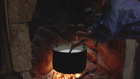 woman stirring milk in cauldron boiling above flames in old fireplace