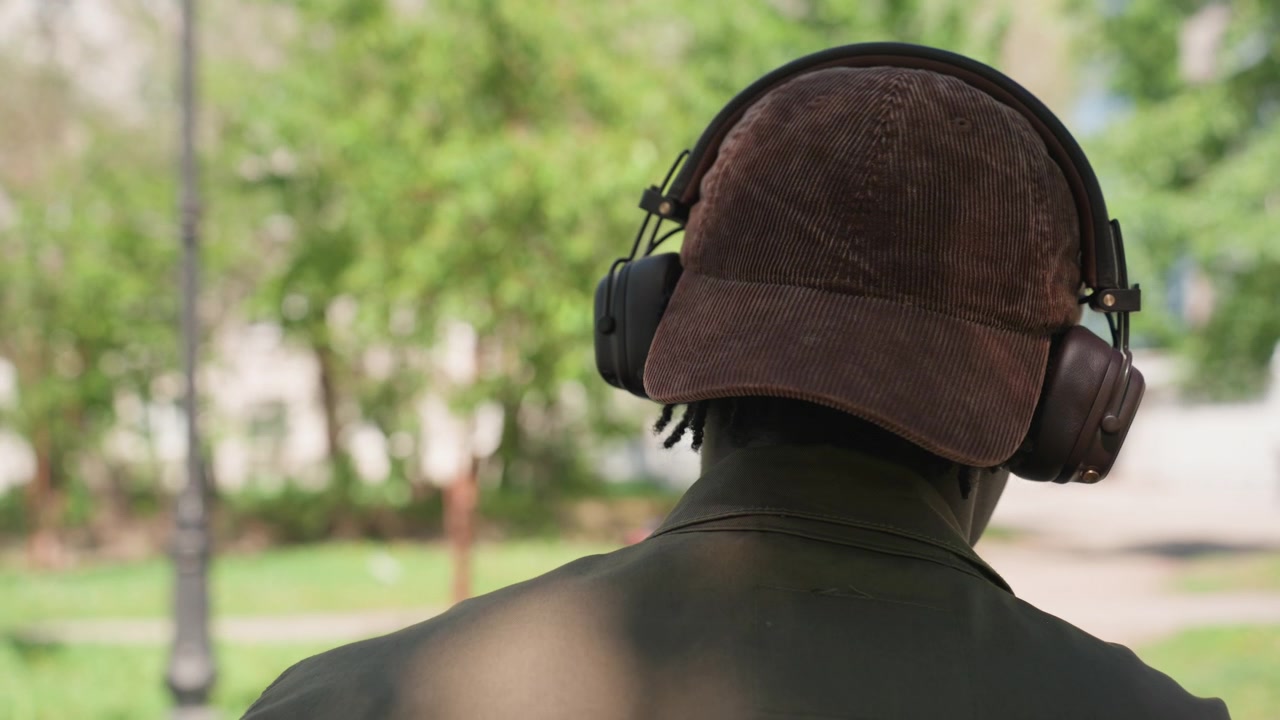 Close up of person wearing headphones and brown cap viewed from behind in outdoor park setting with green trees. person remains stationary suggesting they are listening to music or audio