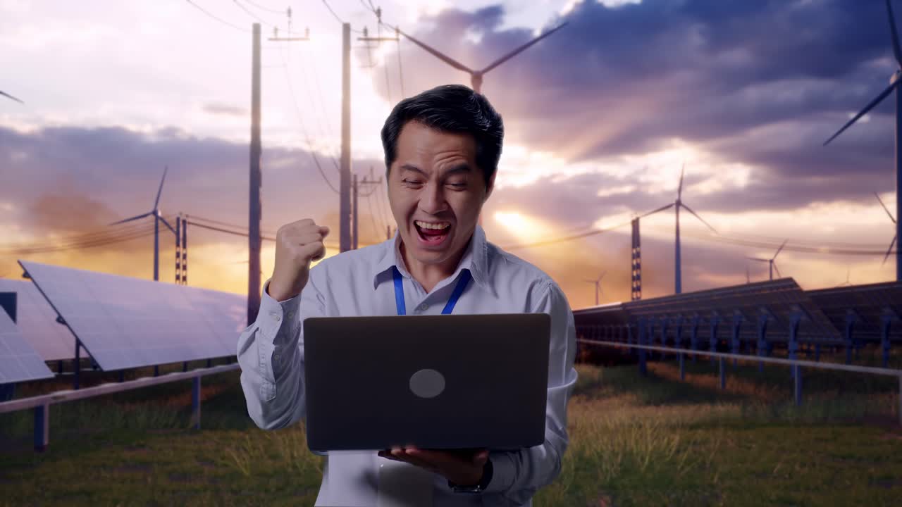 Asian Male Professional Worker Standing With His Laptop With Solar Panel and Wind Turbines, He Raises His Fist Up With Screaming Goal